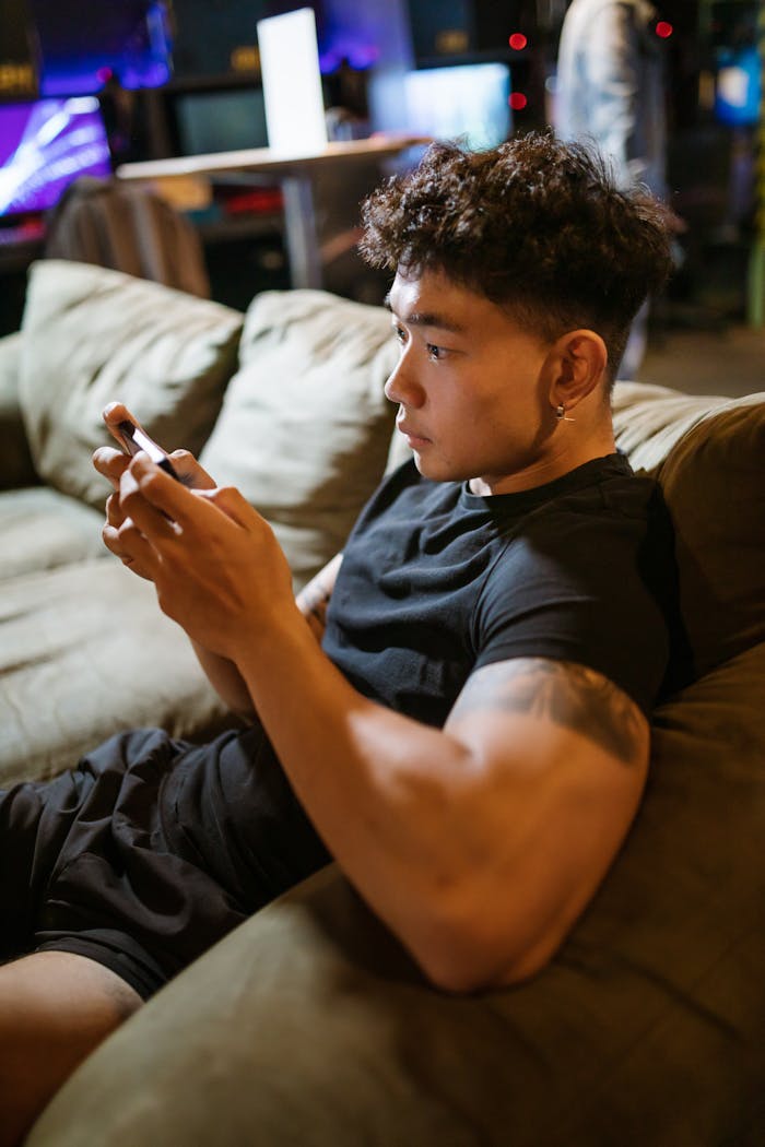 A young man intensely focused on playing a game on his smartphone while lounging on a couch indoors.